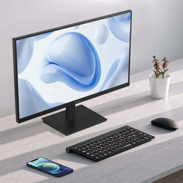 Modern desktop setup with widescreen monitor, wireless keyboard, mouse, smartphone, and small potted plant