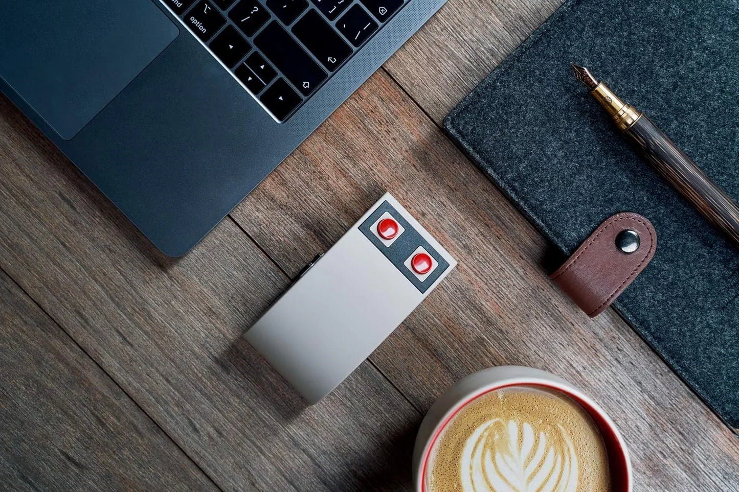 Retro 8-bit gaming mouse with red buttons beside laptop, notebook, pen and coffee on wooden desk.