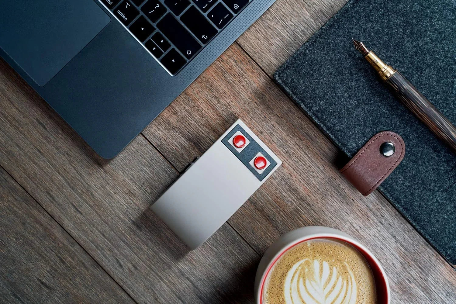 Retro 8-bit gaming mouse with red buttons beside laptop, notebook, pen and coffee on wooden desk.