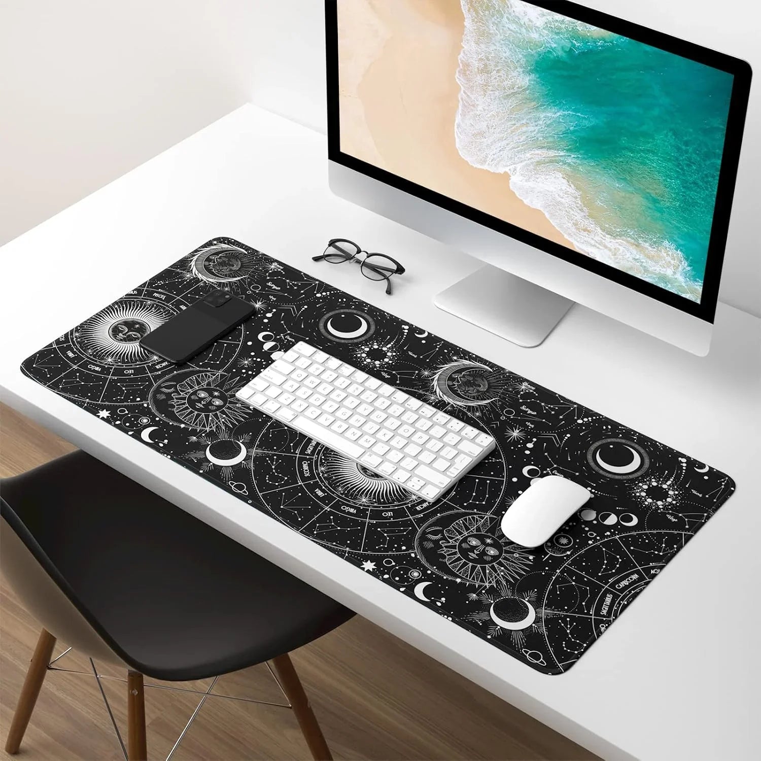Modern white desk setup with large celestial-themed black mouse pad, wireless keyboard and mouse, computer monitor, smartphone, and glasses