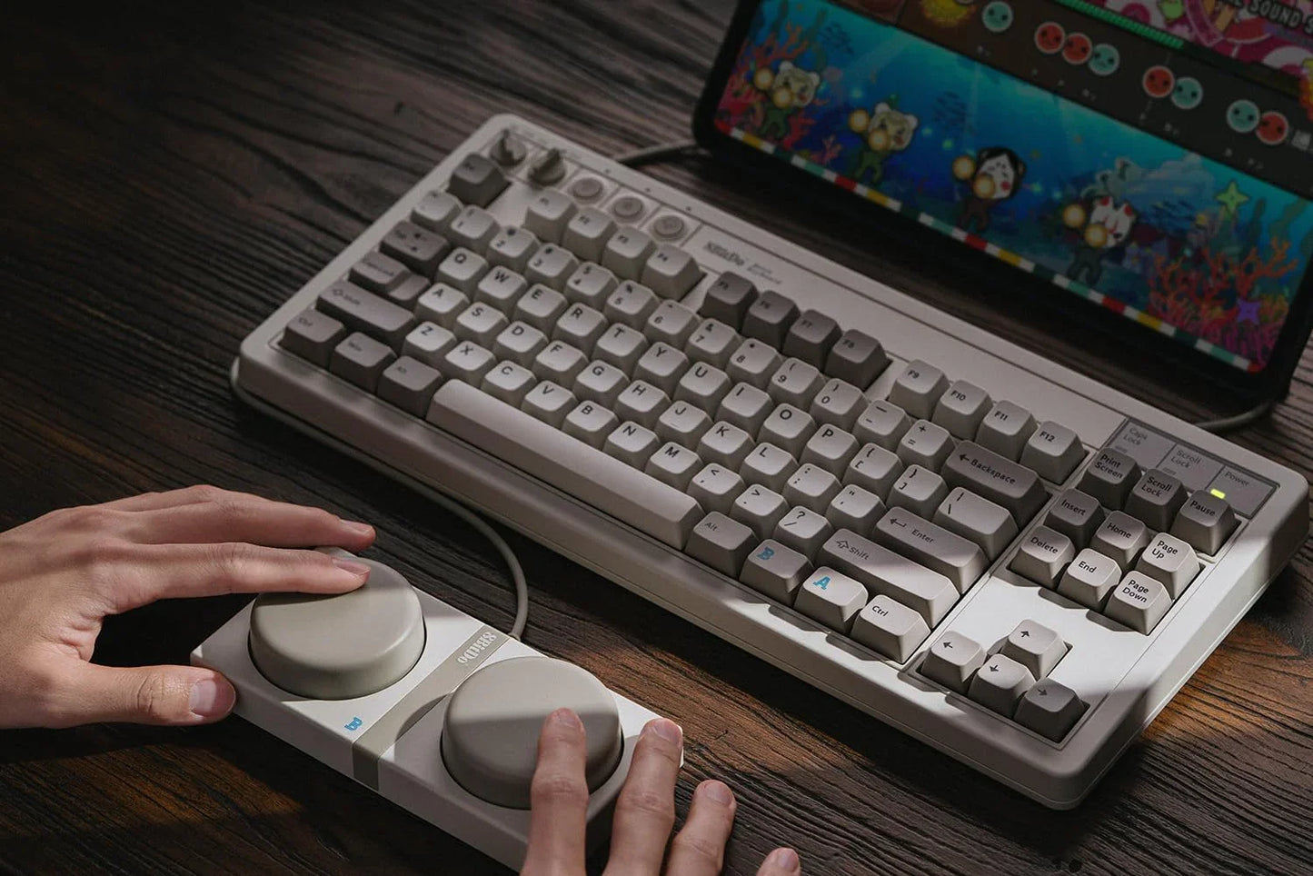 Retro mechanical keyboard and large gaming controller with hands, next to monitor on wooden desk