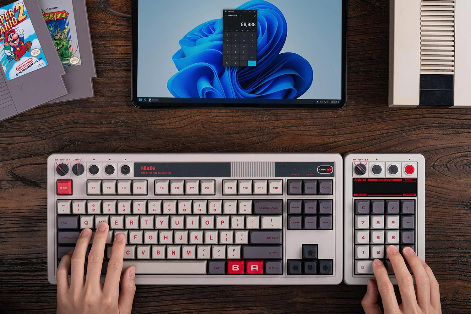 Hands using a retro gaming keyboard and numpad with a vintage computer setup on a wooden desk.