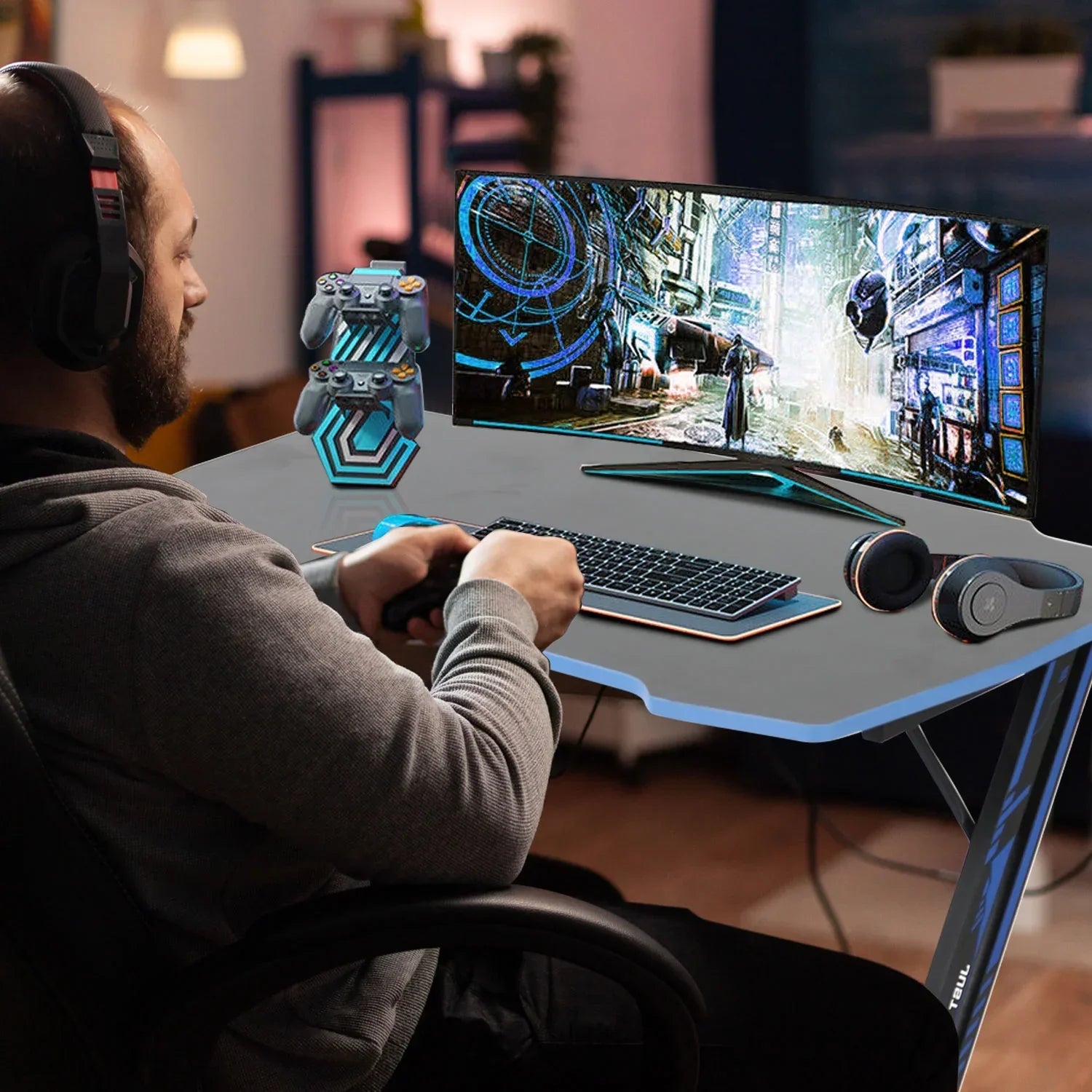 Man playing video games on gaming desk with ultra-wide monitor, wireless keyboard, controllers stand, and headphones