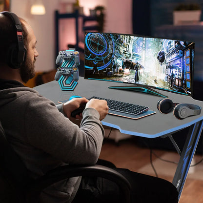 Man playing video games on gaming desk with ultra-wide monitor, wireless keyboard, controllers stand, and headphones