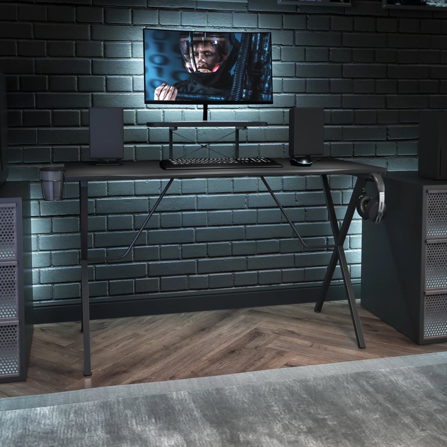 Modern black gaming desk setup with monitor, keyboard, mouse, speakers, and headphones against a gray brick wall