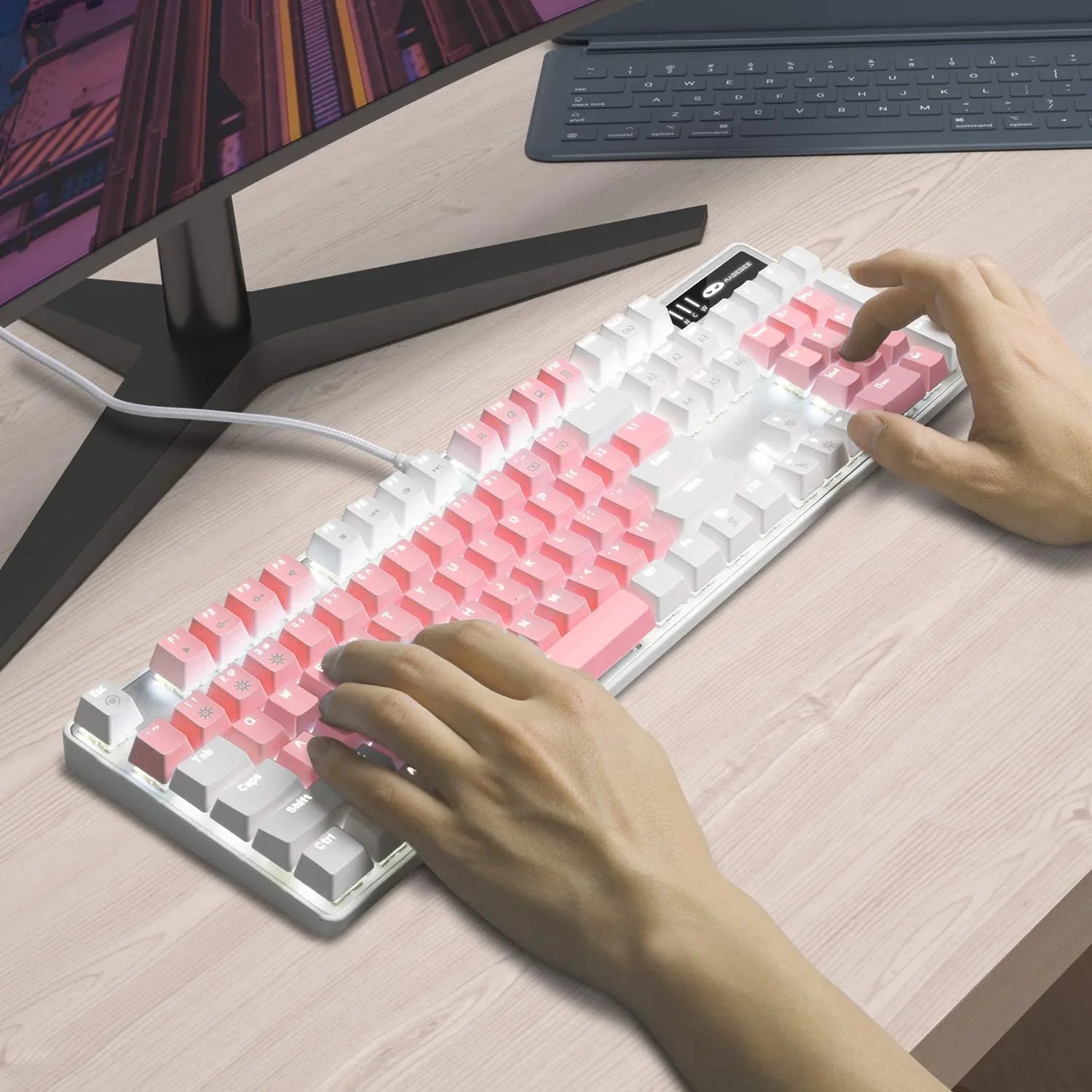 Person typing on white mechanical gaming keyboard with pink and white keycaps on a light wood desk