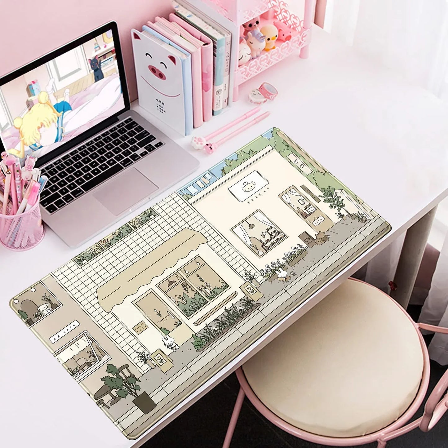 White desk with pink accents featuring a laptop playing anime, cute stationery, and a scenic mouse pad with bakery and café illustration