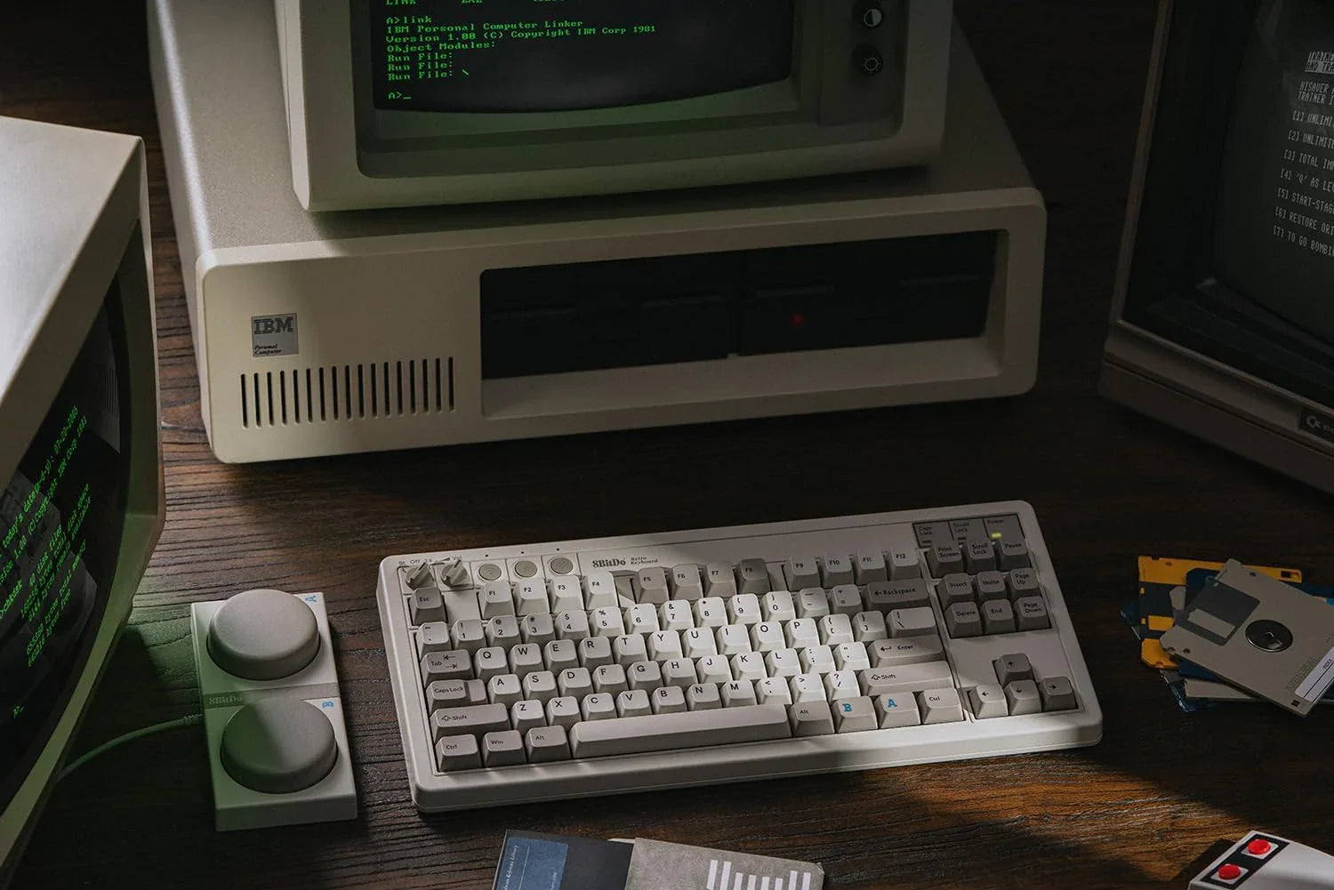Retro IBM computer setup with vintage mechanical keyboard, monitors, floppy disks, and gaming controller.