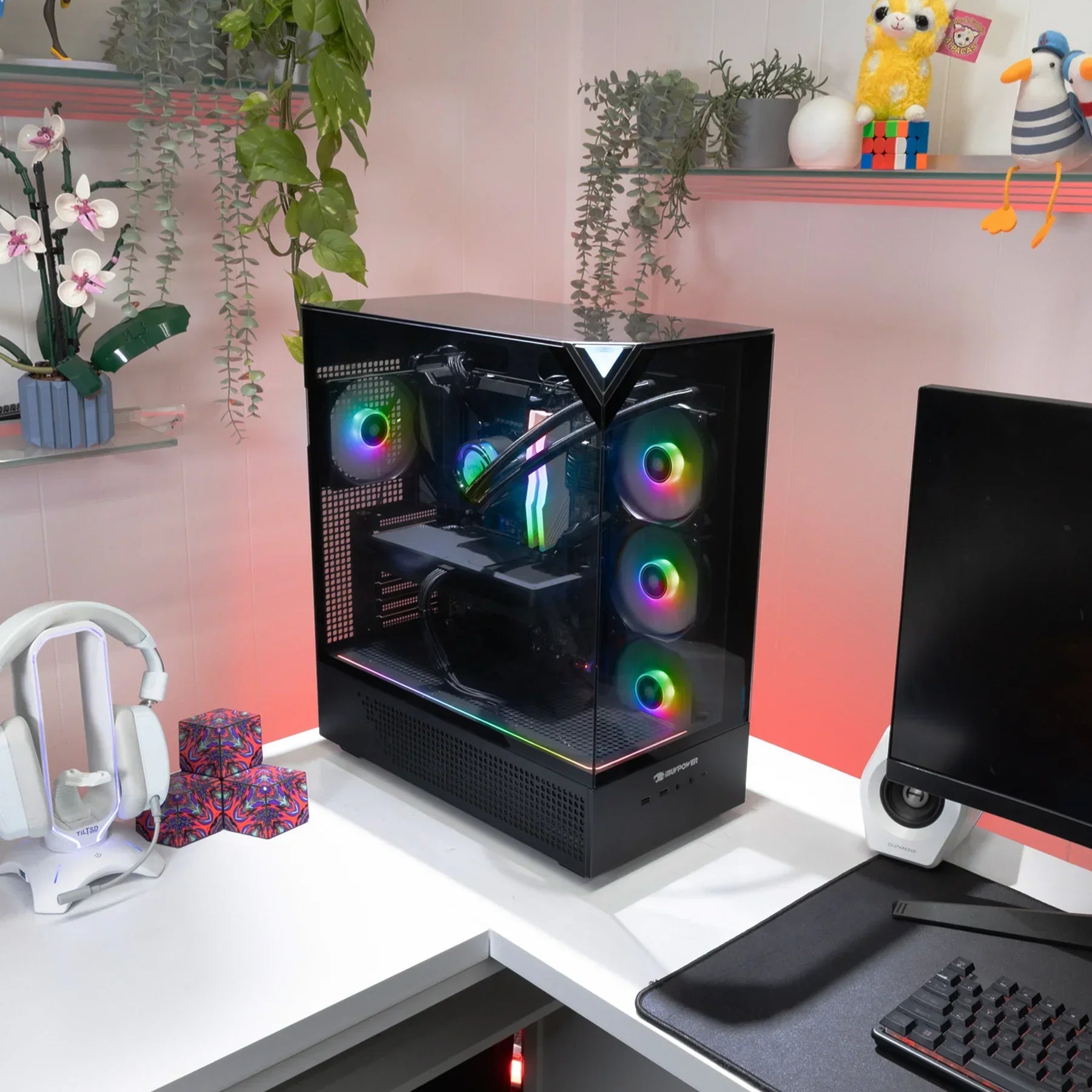Modern gaming PC with RGB fans on white corner desk, surrounded by headphones, monitor, and plants