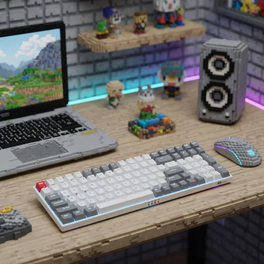 Retro 8-bit gaming desk setup with pixel-style keyboard, mouse, laptop and blocky figures.