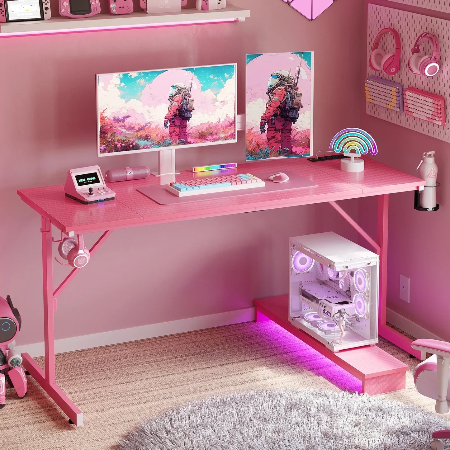 Pink gaming desk setup with dual monitors, colorful keyboard, headset, PC with purple lights, and space-themed astronaut wallpaper