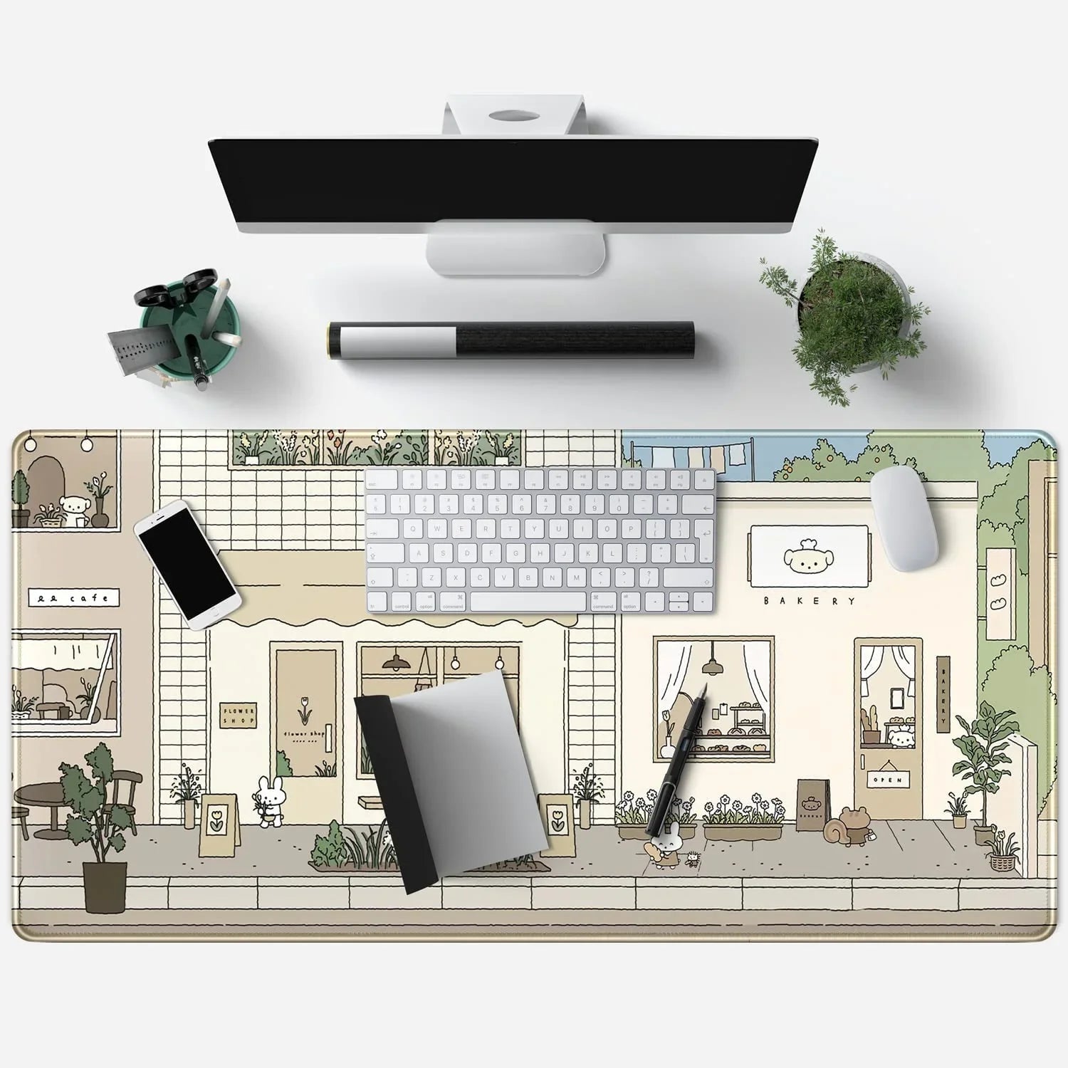 Top view of a clean desk with a monitor, keyboard, mouse, cute cartoon town-themed desk mat, smartphone, notebook, pen, and a small plant