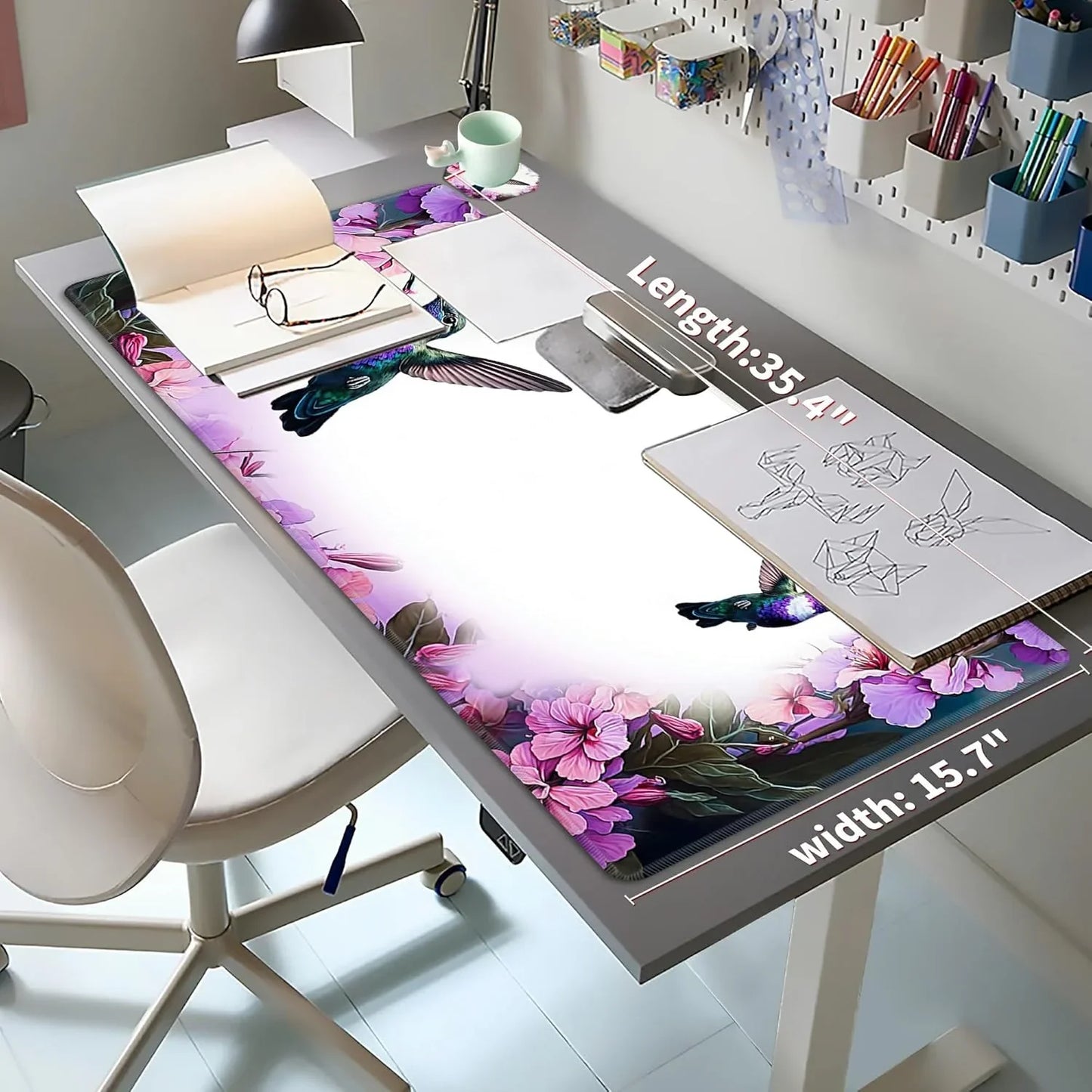 Desk setup with floral and hummingbird-themed large mouse pad, sketchbook, glasses, and stationery on gray table with white chair