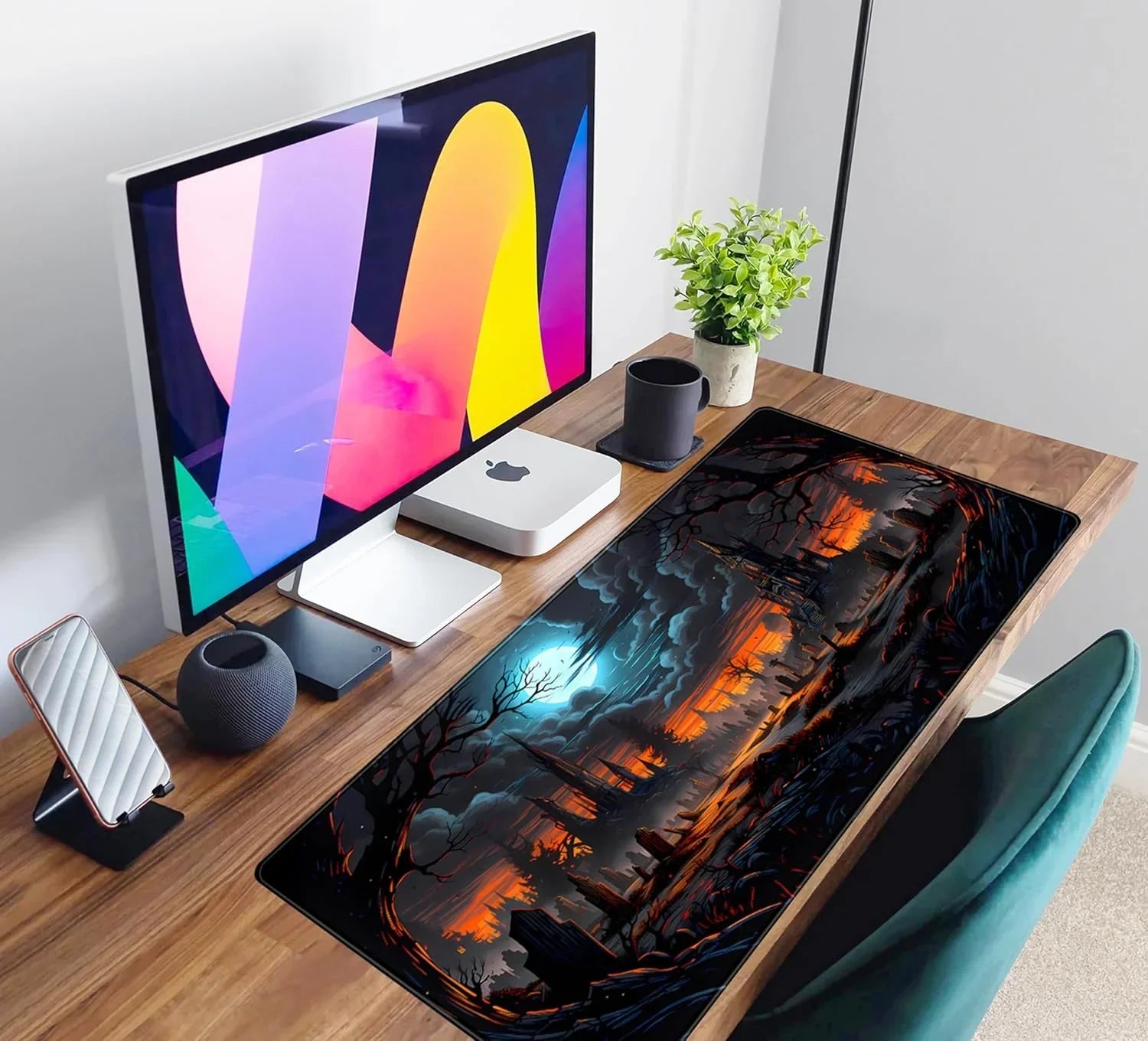 Modern wooden desk setup with Apple desktop, colorful monitor, black mug, green plant, and Halloween-themed gaming mouse pad
