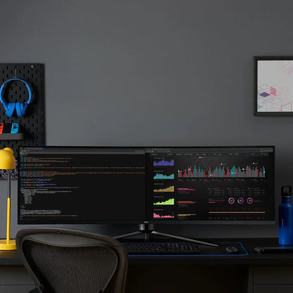 Ultra-wide curved monitor on black desk showing coding and data charts, with yellow lamp, blue headphones, and gaming gear