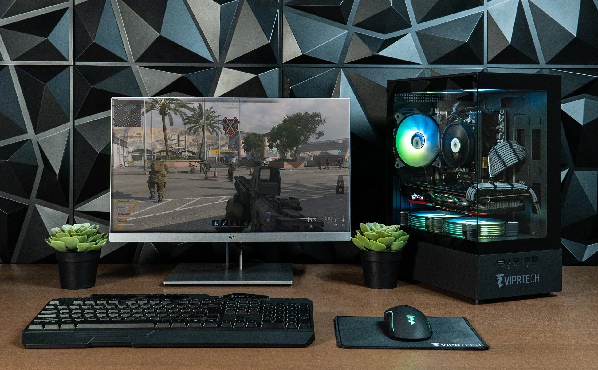 Gaming setup with RGB-lit VIPRTECH PC, HP monitor displaying first-person shooter game, mechanical keyboard, mouse on pad, and two small green plants