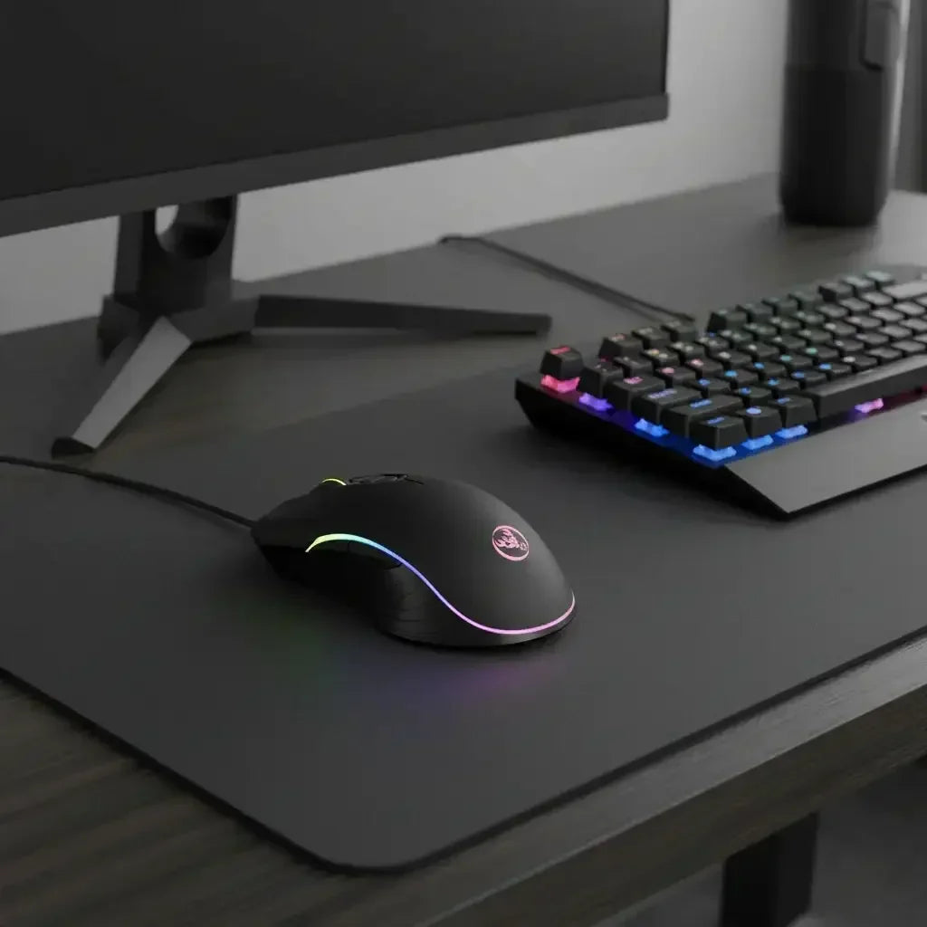 Black wired gaming mouse with RGB lighting on black desk mat beside RGB mechanical keyboard and monitor