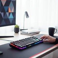 RGB mechanical keyboard on desk with hand typing, black mug, plant, modern monitor, and lamp