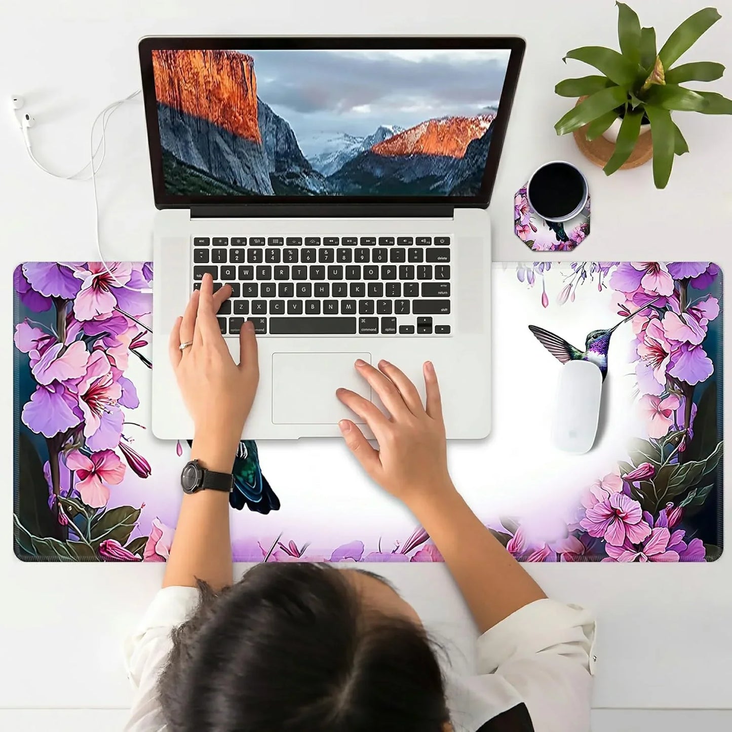 Overhead view of a person using a laptop on a floral-printed extended gaming mouse pad with hummingbird design, a plant, and a coffee cup on white desk