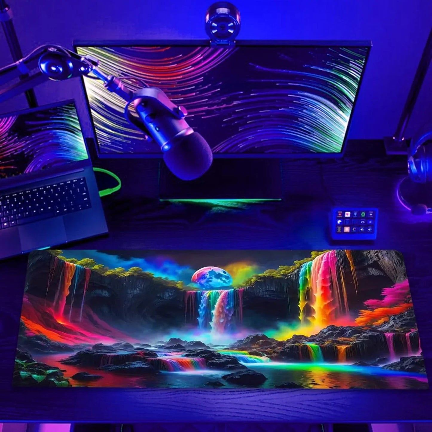 Gaming desk setup with RGB lighting, widescreen monitor, laptop, microphone, and vibrant rainbow waterfall mousepad