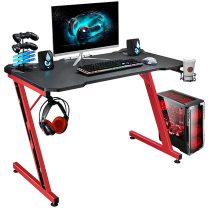 Modern gaming desk with carbon fiber surface, red metal frame, RGB keyboard, gaming mouse, dual speakers, dual controllers, headset hook, cup holder, and PC tower with red cooling fans