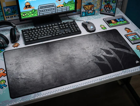 Large black gaming mouse pad on retro 8-bit themed desk setup with keyboard, mouse, headset, and pixel art decor.