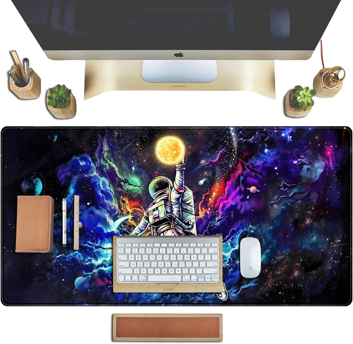 Desk setup with colorful astronaut galaxy desk mat, Apple keyboard and mouse, wooden monitor stand, desk accessories, and succulent plants