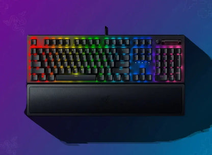 Black gaming keyboard with RGB backlighting and wrist rest on a purple-blue gradient background